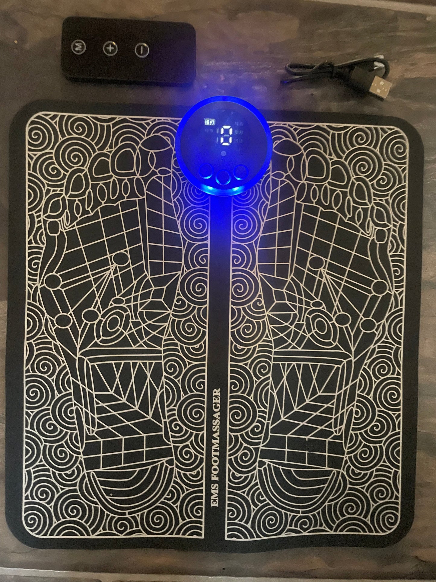 EMS fotmassasjematte med digitale kontroller, avbildet på etgulv sammen med en fjernkontroll og en USB-ladekabel. Matten har et dekorativt mønster med fotrefleksologi-design og en opplyst LED-skjerm i midten.