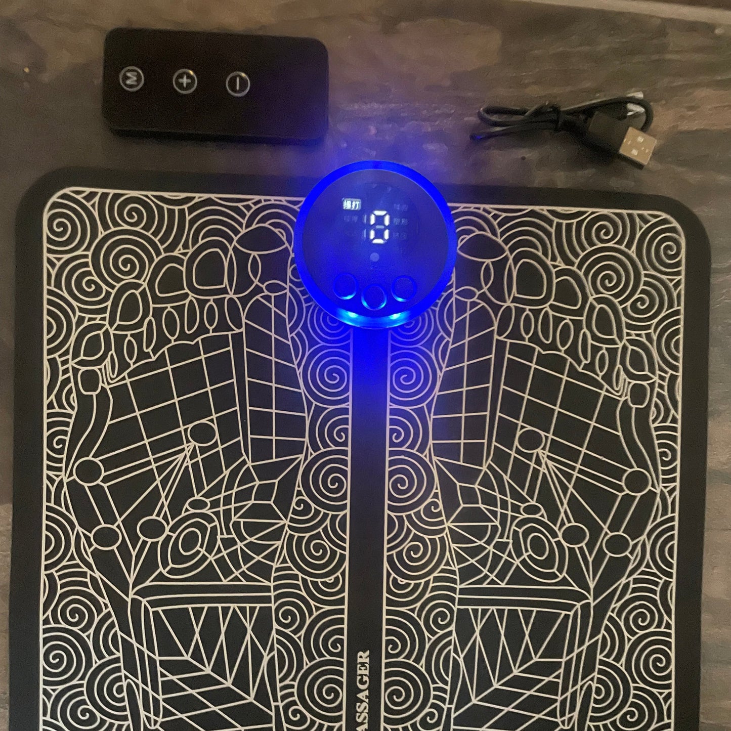 EMS fotmassasjematte med digitale kontroller, avbildet på etgulv sammen med en fjernkontroll og en USB-ladekabel. Matten har et dekorativt mønster med fotrefleksologi-design og en opplyst LED-skjerm i midten.