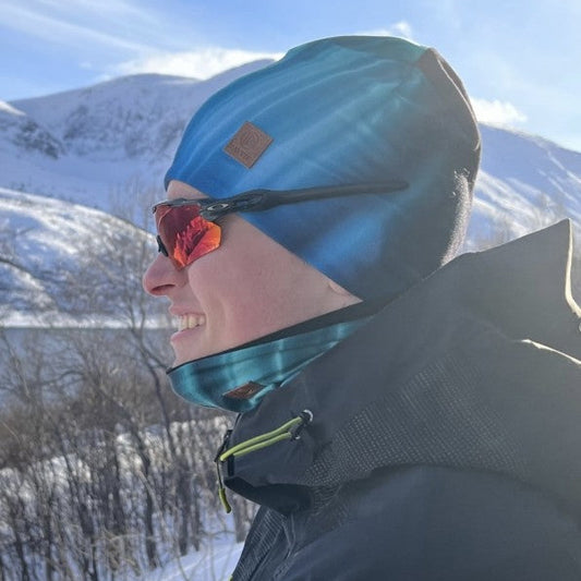 Person med solbriller og blå Davvin® Northern Lights lue og hals, smilende i vinterlandskap med snødekte fjell i bakgrunnen.