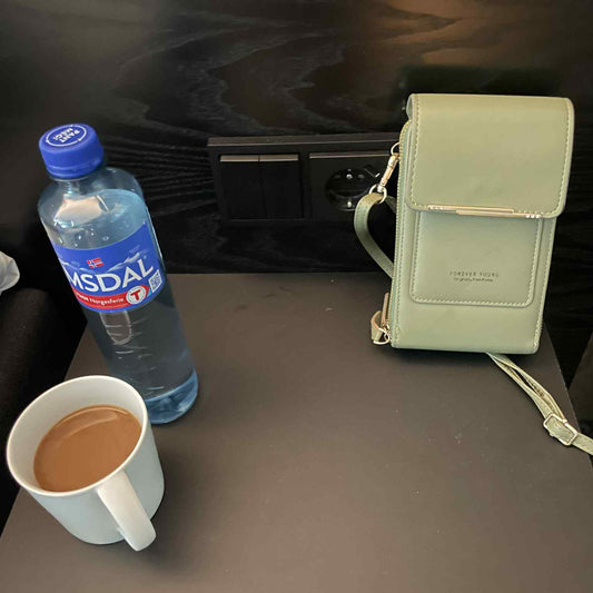 Lys grønn mobilveske med teksten "Forever Young" plassert ved siden av en kopp kaffe og en flaske Imsdal vann på et mørkt bord med stikkontakt i bakgrunnen.