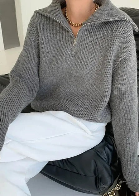En grå genser med glidelås. Genseren har en klassisk passform og ribbestrikkede detaljer ved ermer og hals. Glidelåsen går fra halsen ned til brystet, noe som gir mulighet for justering av kragen. Stoffet er mykt og behagelig, ideelt for både hverdagsbruk og avslappende øyeblikk.