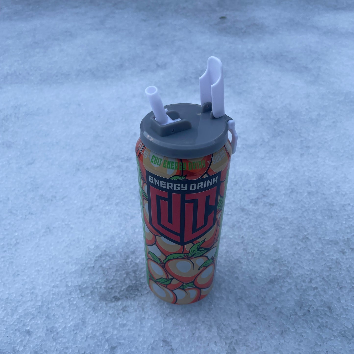 Grått bruslokk med integrert sugerør festet på en energidrikkboks, plassert på snø.