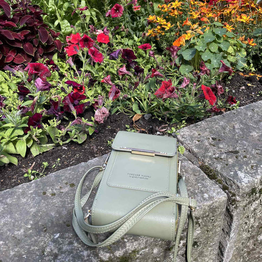 Lysegrønn liten skulderveske med teksten "Forever Young" plassert på en steinmur foran et fargerikt blomsterbed med røde, oransje, lilla og rosa blomster.