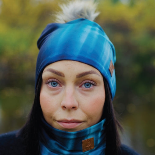 Person med blå Davvin® Northern Lights lue med pelsdusk og tilhørende hals. Naturen er uskarp i bakgrunnen.