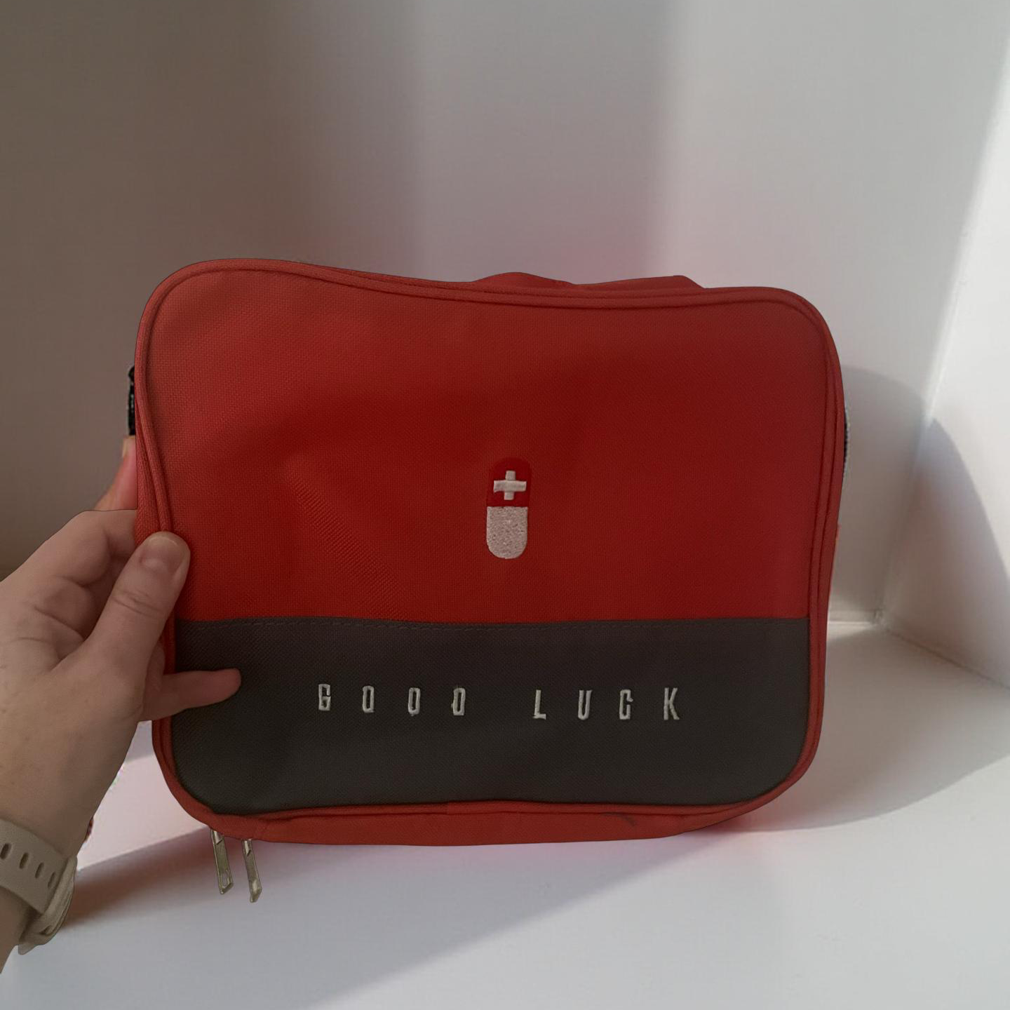 Rød førstehjelpsveske med teksten 'GOOD LUCK' og et ikon av en pille med et hvitt kors på. En hånd holder vesken mot en lys bakgrunn.