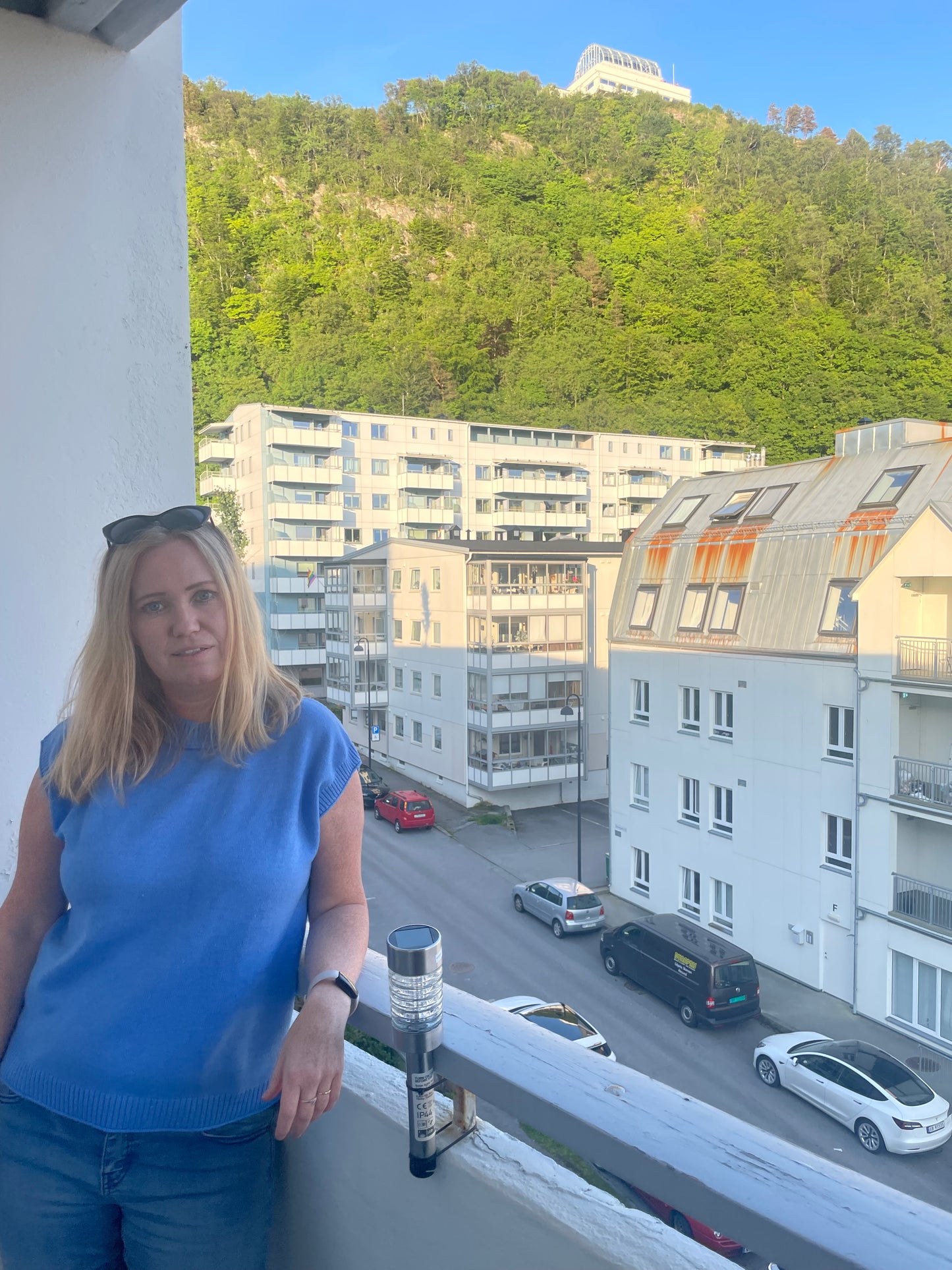 Kvinne med langt lyst hår og blå ermeløs strikket topp står avslappet på en balkong i solnedgangslys. Hun ser mot kameraet med et svakt smil. Bak henne ses en grønn skogkledd ås med en utsiktsbygning på toppen, og flere moderne bygårder og parkerte biler i gaten under.