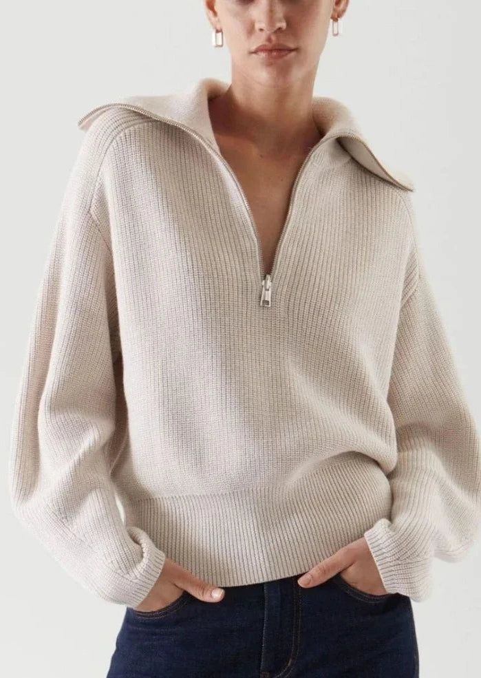 En beige genser med glidelås. Genseren har en klassisk passform og ribbestrikkede detaljer ved ermer og hals. Glidelåsen går fra halsen ned til brystet, noe som gir mulighet for justering av kragen. Stoffet er mykt og behagelig, ideelt for både hverdagsbruk og avslappende øyeblikk.